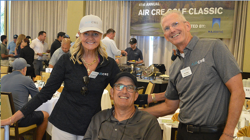 Three people smiling for a photo during a post-golf dinner banquet with people in the background and a sign that says AIR CRE Golf Classic
